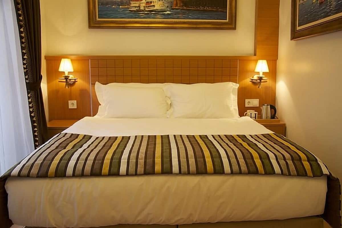 A hotel room with a large bed, two bedside lamps, a painting above the bed, and a nightstand with a coffee pot.