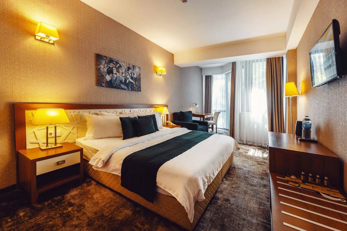 A hotel room with a large bed, bedside lamps, a desk with a chair, a television, and a painting on the wall.