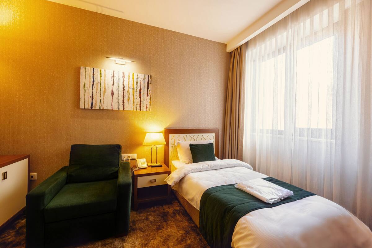 A hotel room with a bed, a chair, a nightstand, and a painting on the wall.