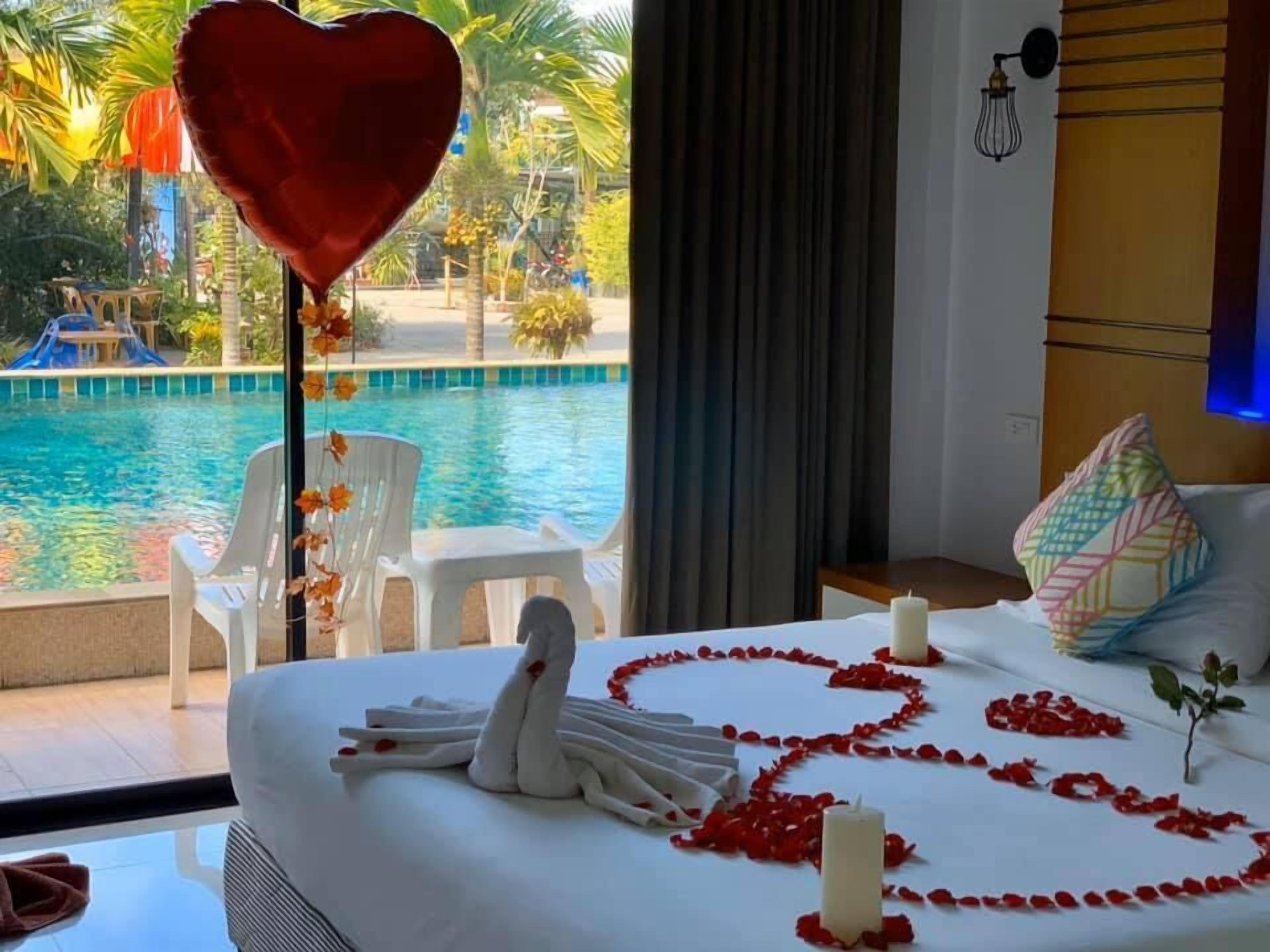 Habitación de hotel con una cama decorada con un cisne y pétalos de rosa, un globo con forma de corazón y vistas a una piscina y palmeras.