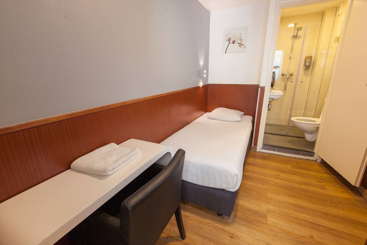 Una habitación de hotel pequeña y funcional con dos camas individuales, un banco de madera, un lavamanos blanco y una cabina de ducha de vidrio.