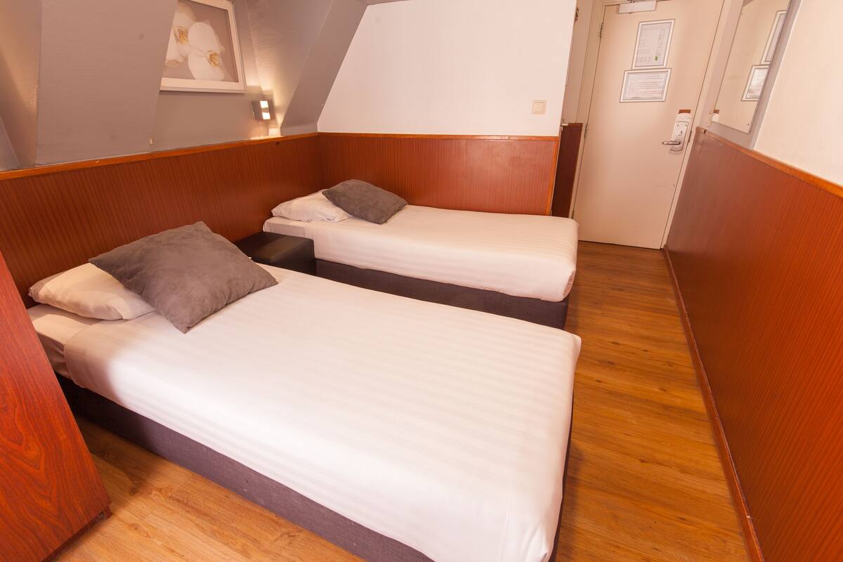 Una habitación compacta con dos camas individuales, cada una con una almohada y una mesita de noche pequeña. La habitación tiene paneles de madera y una puerta con un espejo y un aviso.
