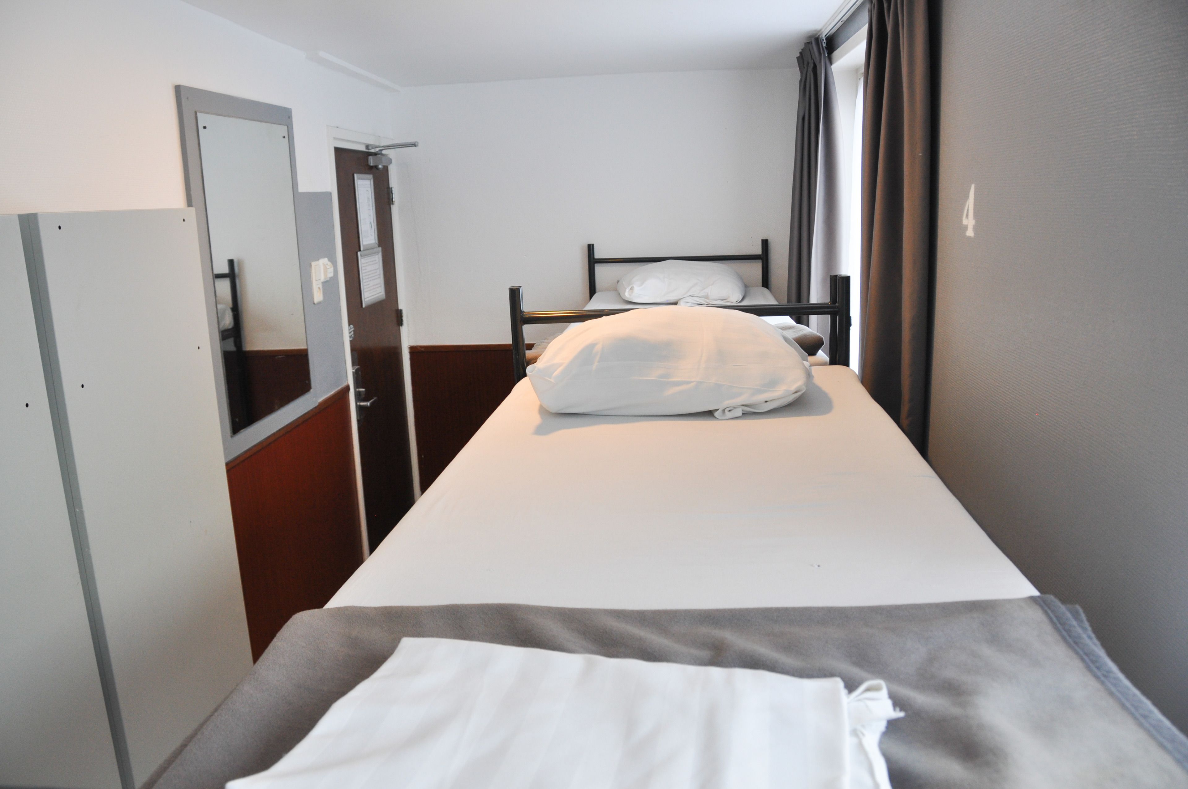 A single bed with a white mattress and pillow, a wooden headboard, a small bedside table, and a door to the left.