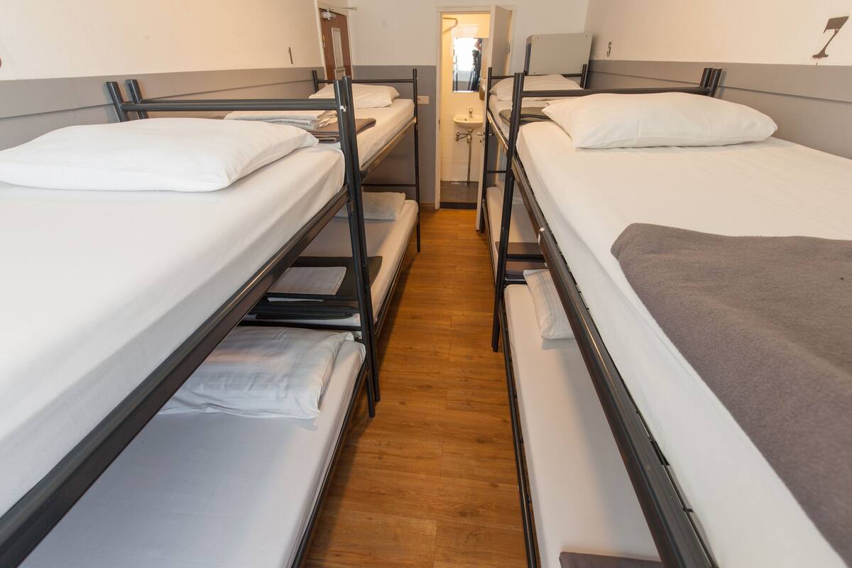 Habitación tipo dormitorio con literas, suelo de madera y un baño pequeño con lavabo y espejo.