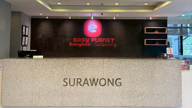 A reception desk with a sign for Easy Planet Bangkok and Surawong, a tiled floor, and a ceiling with recessed lighting.