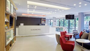 A modern reception area with a reception desk labeled "SURAWONG," a bookshelf, and seating arrangements including red chairs and tables.