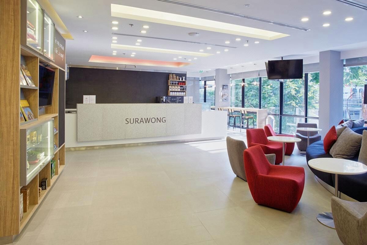 Zona de recepción con mostrador identificado como 'SURAWONG', una estantería y disposición de asientos que incluyen sillas y mesas rojas.