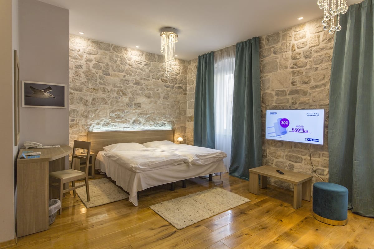 Quarto com parede de pedra, cama, escrivaninha, cadeira, televisão e mesa pequena.