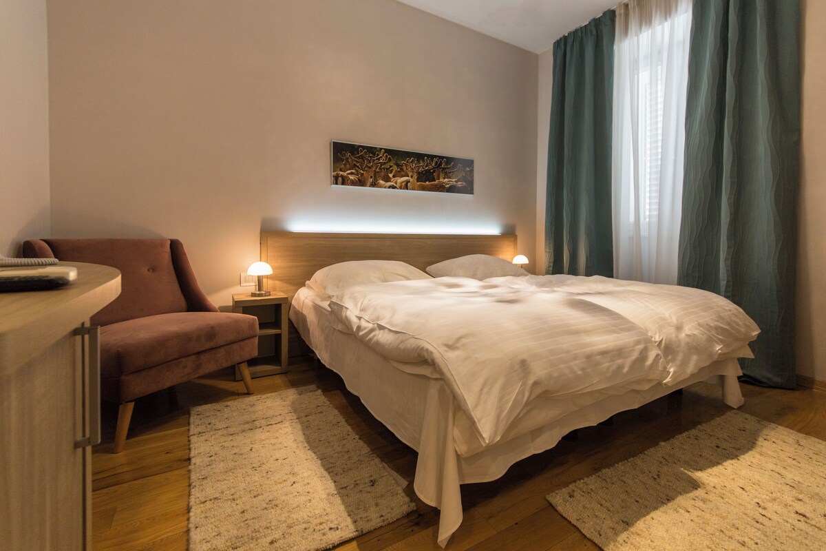 A hotel room with a bed, a chair, a nightstand, and a painting on the wall.