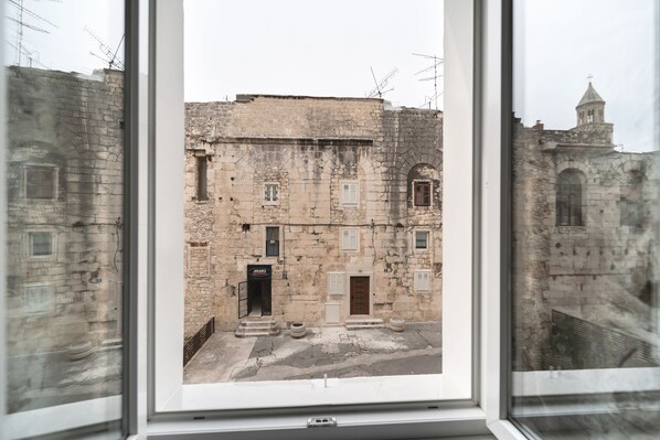 A view through a window of a stone building with a door labeled "MAZZA," a small courtyard, and a distant church tower. - Tifani Luxury Rooms (Split)