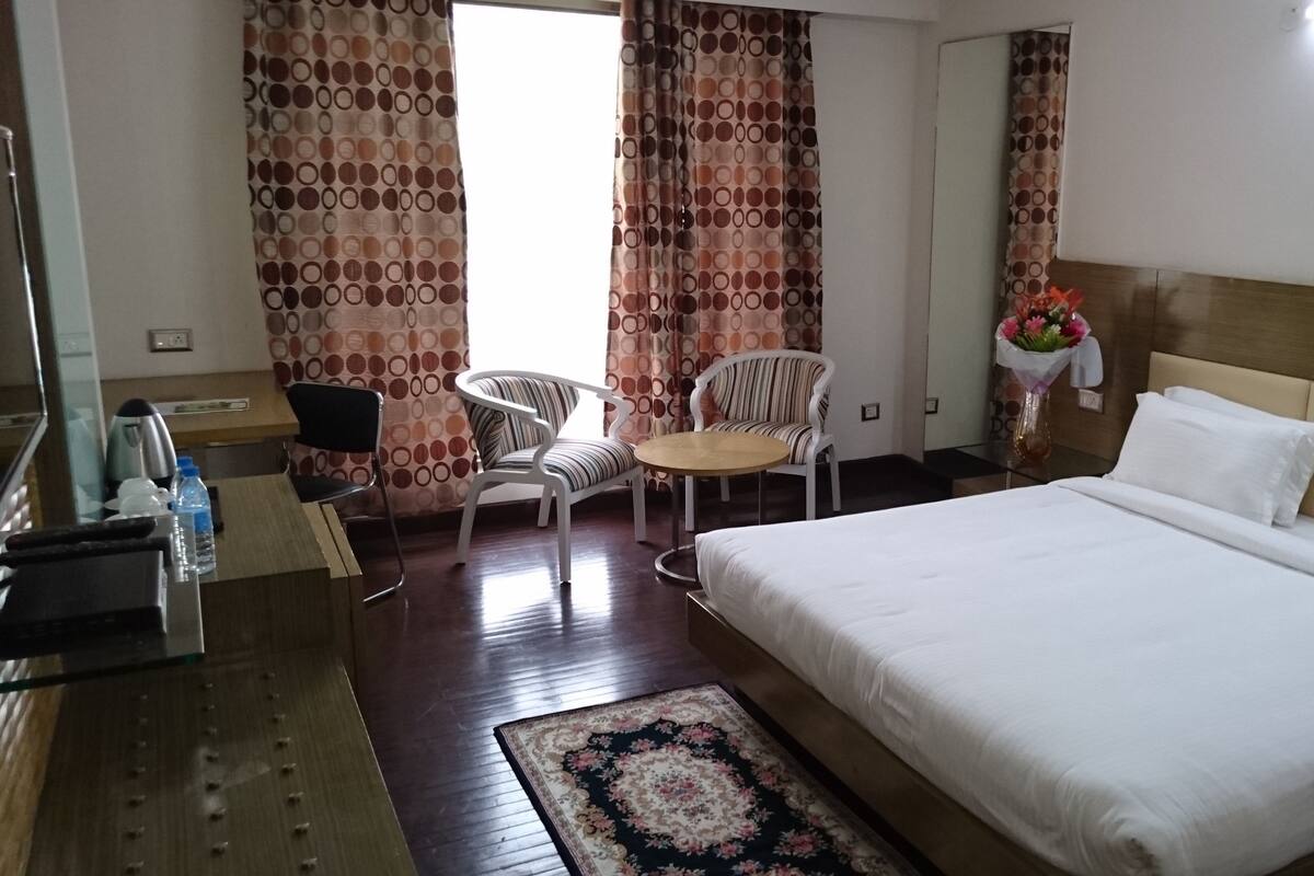 A hotel room with a bed, two chairs, a small table, a coffee machine, and a vase with flowers.