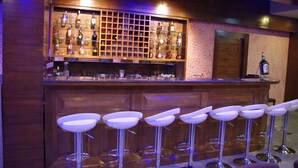Een bar met een houten toonbank, meerdere flessenplanken en witte barstoelen.
