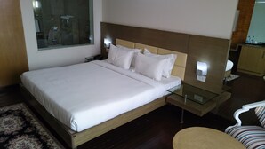 Een net opgemaakt bed met witte lakens, een nachtkastje met een lamp, en een klein rond tafeltje met een stoel.