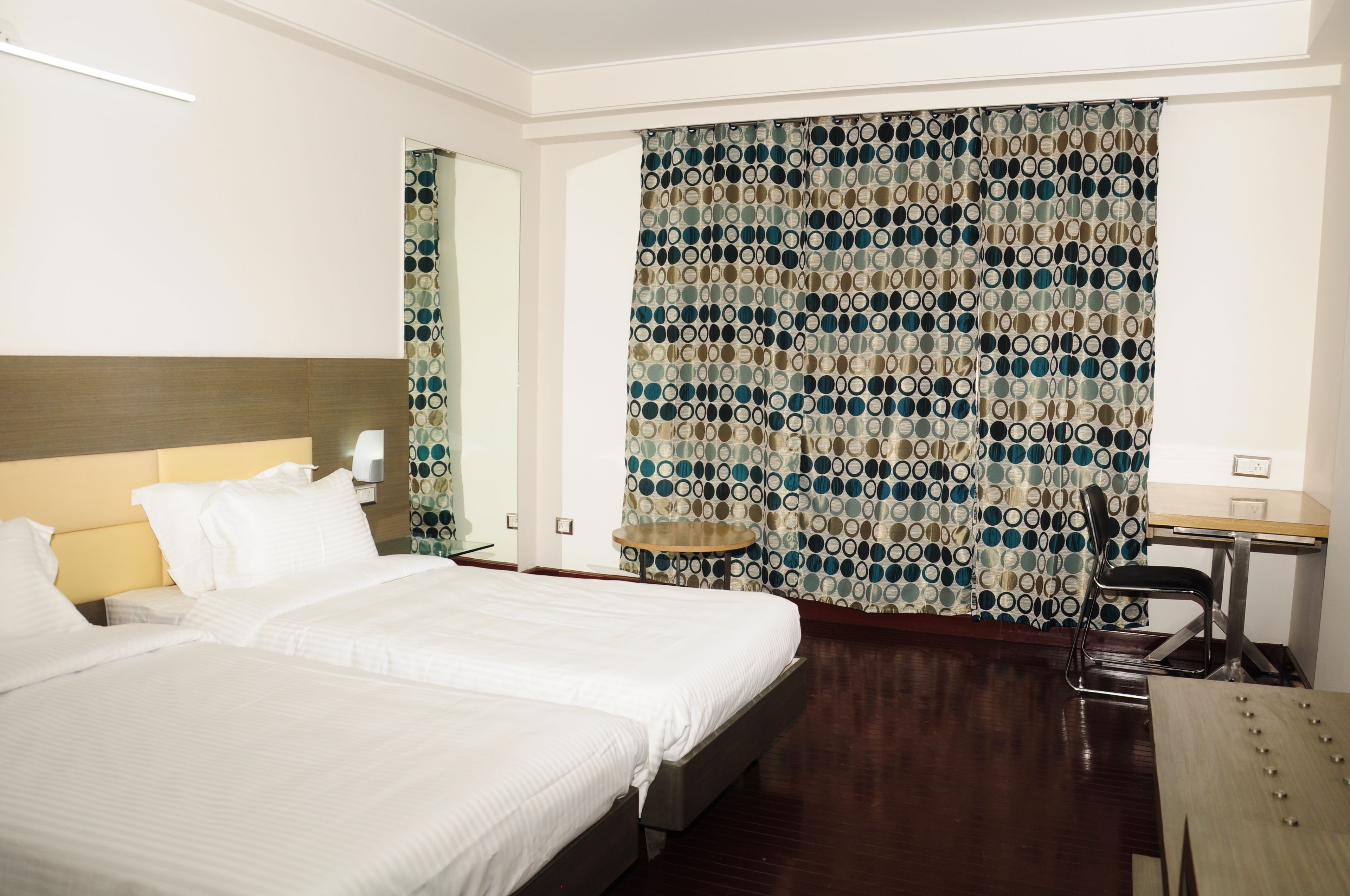 A hotel room with two beds, a desk, a chair, and a patterned curtain.