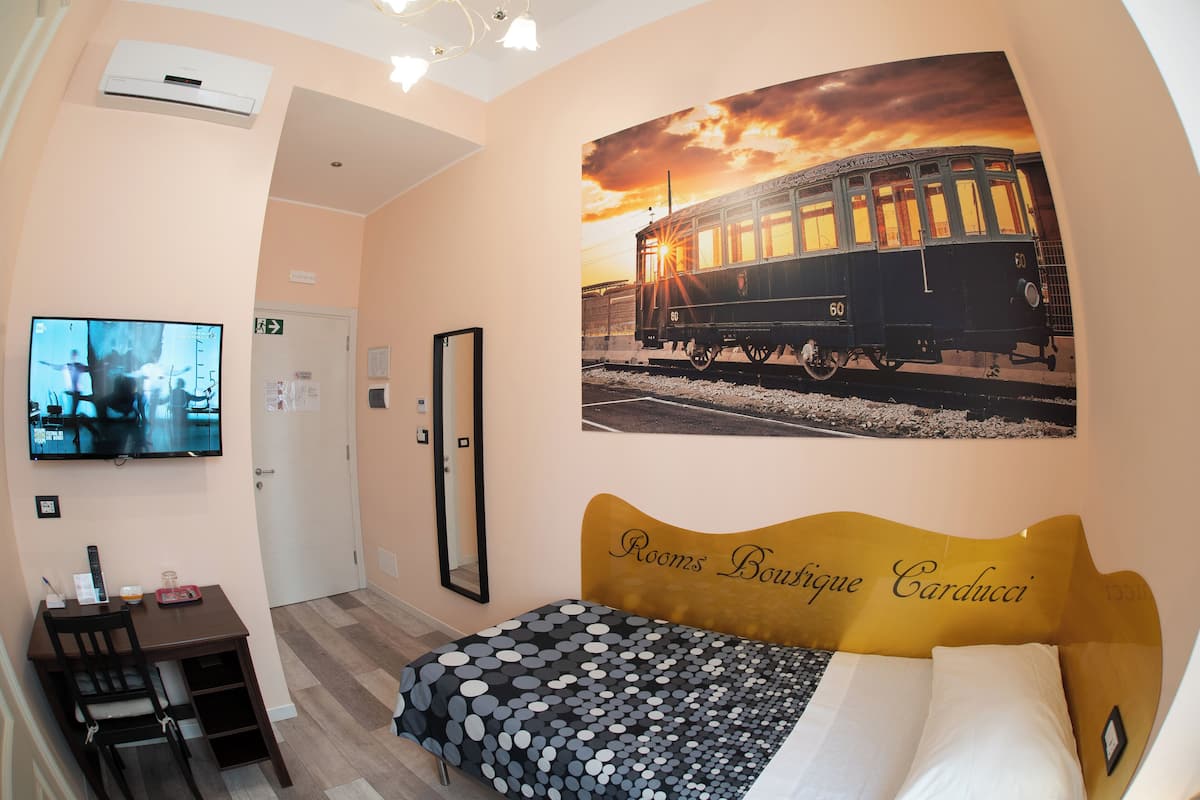 Una habitación de hotel con una cama, un televisor en la pared, un escritorio con una silla y una gran fotografía de un tren en la pared. Se identifica como Rooms Boutique Carducci.