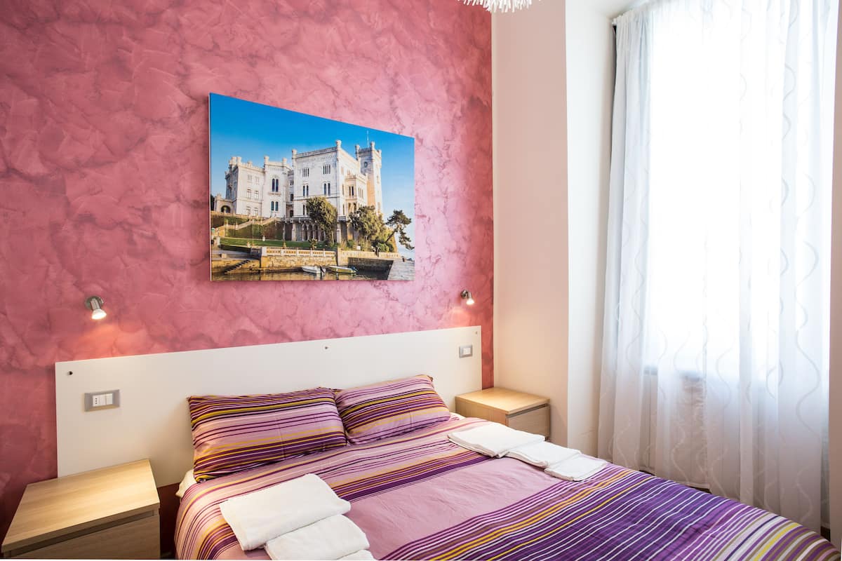 Un dormitorio con una cama, dos mesitas de noche, una lámpara de noche, un cuadro de un castillo y una ventana con cortinas.