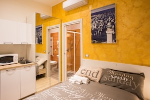 Una habitación de hotel compacta con kitchenette, ducha y cama con almohadones decorativos.