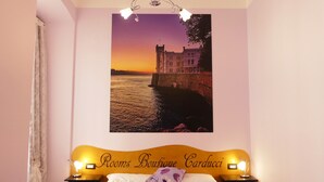 A bedroom with a bed, two bedside tables, a nightstand, a wall-mounted lamp, and a framed picture of a castle by the water. - Rooms Boutique Carducci (Trieste)