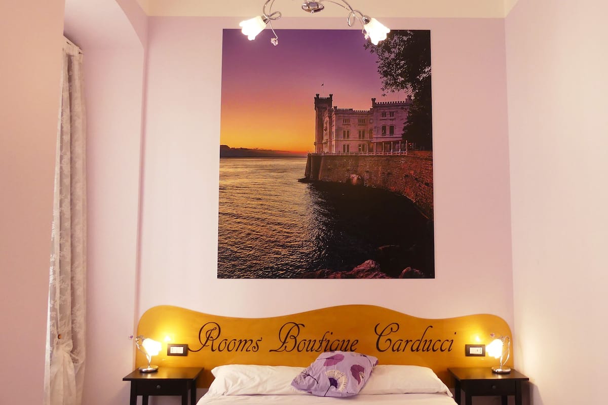 Un dormitorio con una cama, dos mesitas de noche, una lámpara de pared y un cuadro enmarcado que muestra un castillo junto al agua.