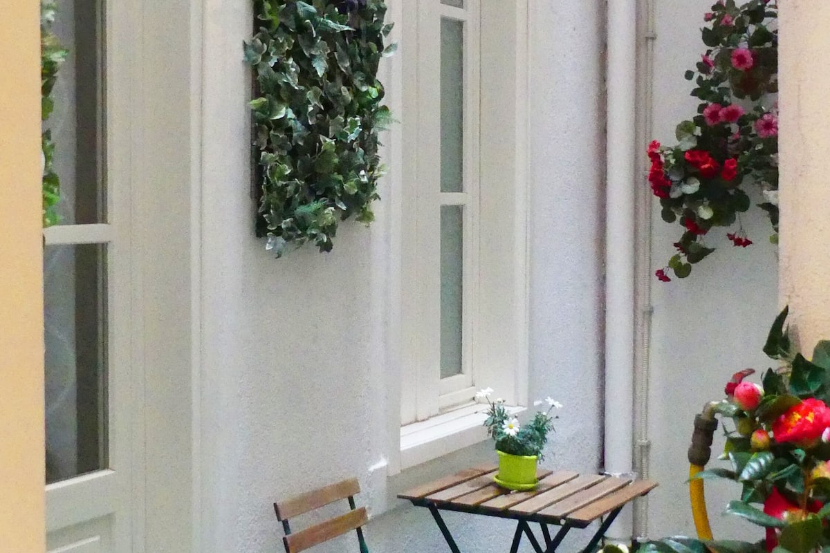 Un pequeño espacio al aire libre con mesa y sillas de madera, rodeado de muros con ventanas verticales y plantas en macetas.