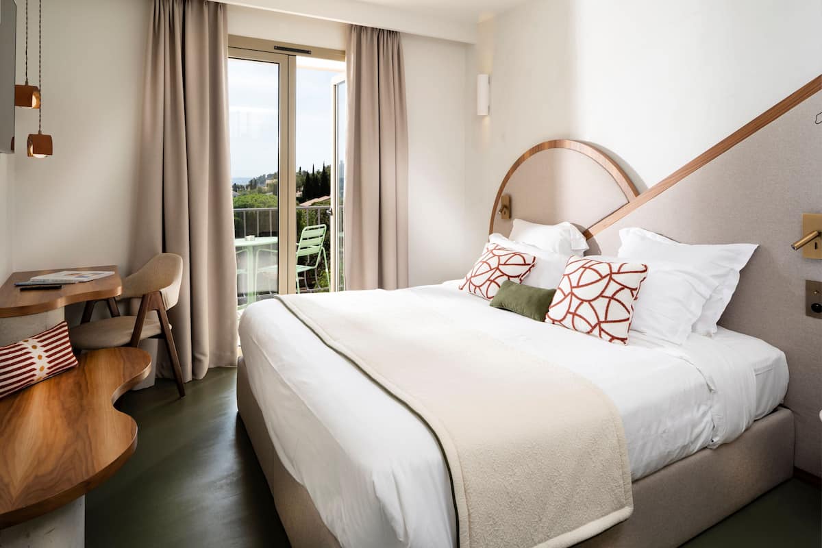 A hotel room with a large bed, a wooden desk, a chair, and a balcony with a view.
