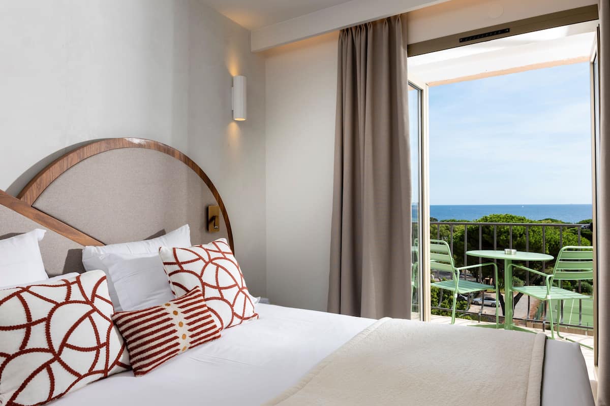 A hotel room with a bed, a view of the outdoors, and a balcony with chairs.