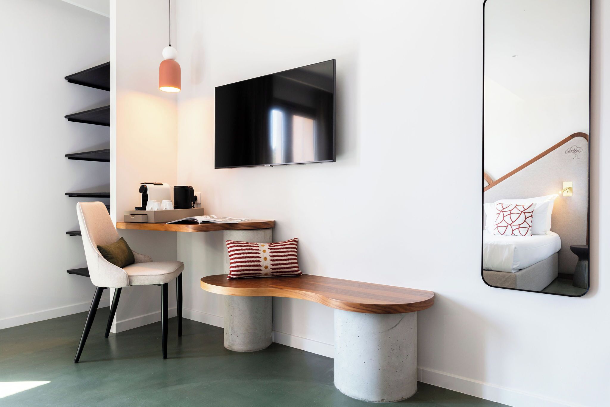 A modern room with a flat-screen TV mounted on the wall, a wooden bench, a chair, a desk with a computer, and a hanging pendant light.