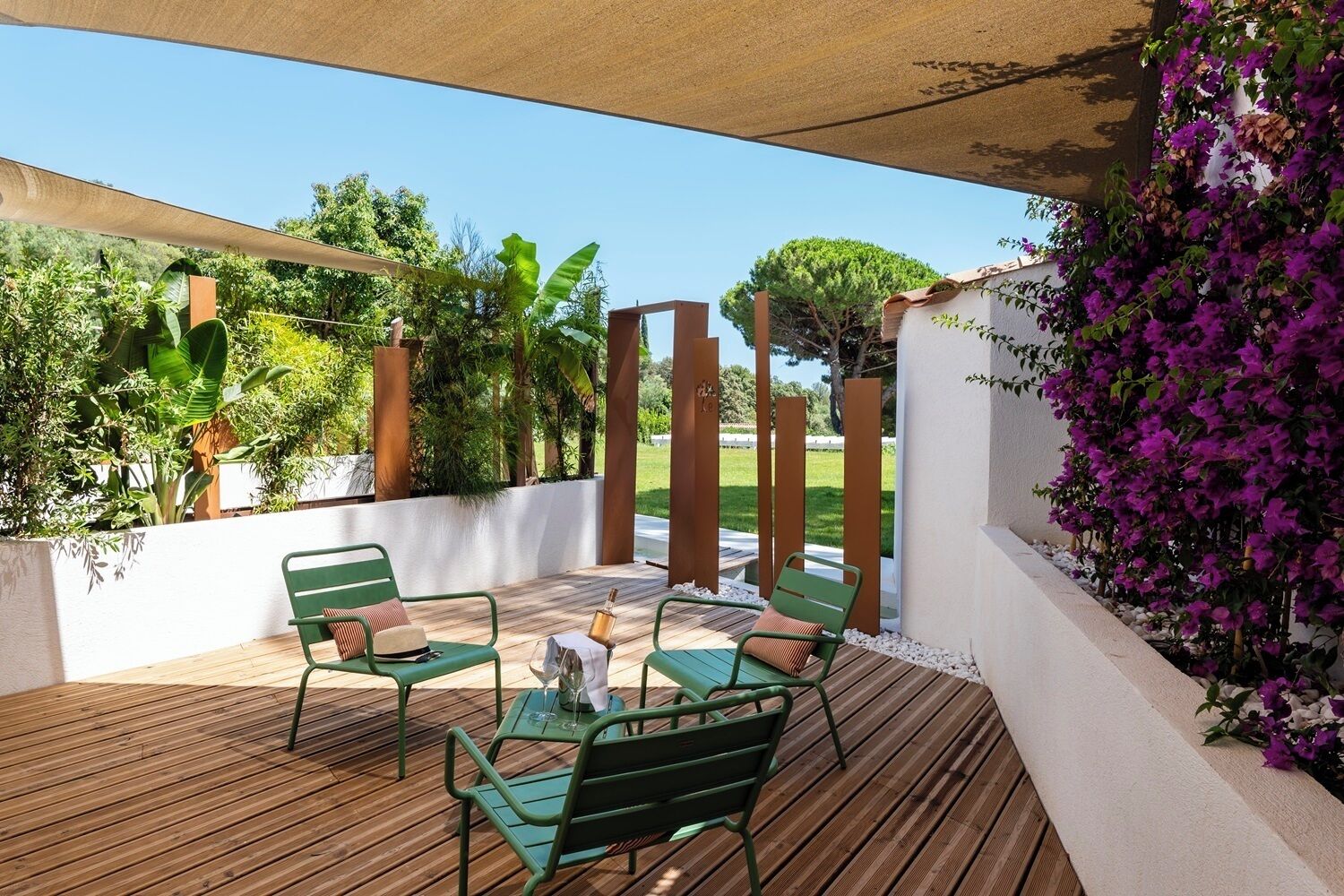 An outdoor patio with wooden flooring, green metal chairs, a table with a bottle and glass, and a view of greenery and trees.
