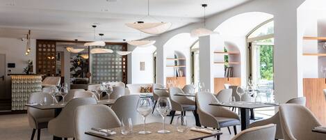 A modern restaurant interior with round tables, beige chairs, and hanging pendant lights.