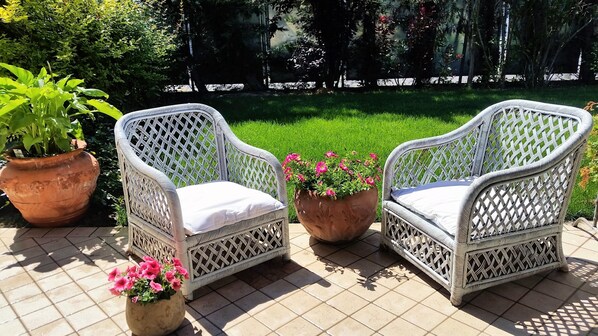 Dos sillas de mimbre con cojines en un patio empedrado, rodeado de plantas en macetas y un césped verde exuberante.