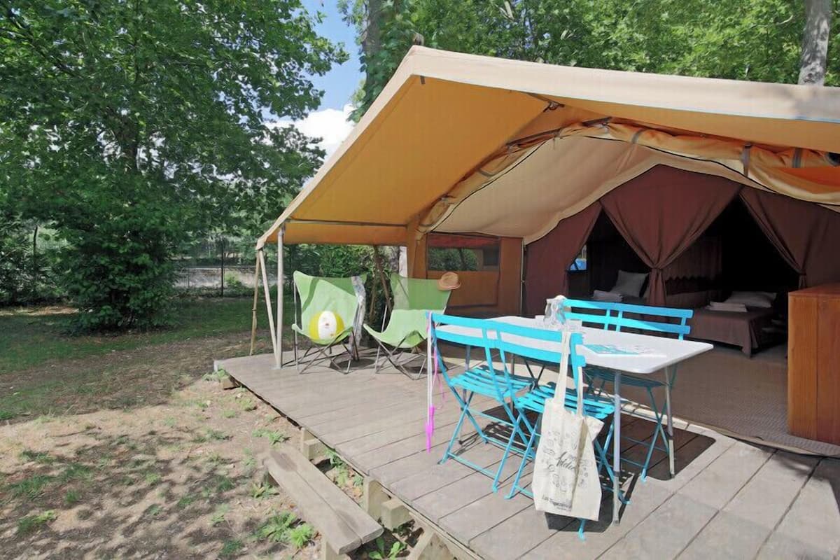 Acomodação em tenda com mesa e cadeiras, um banco e uma pequena mesa com uma garrafa.