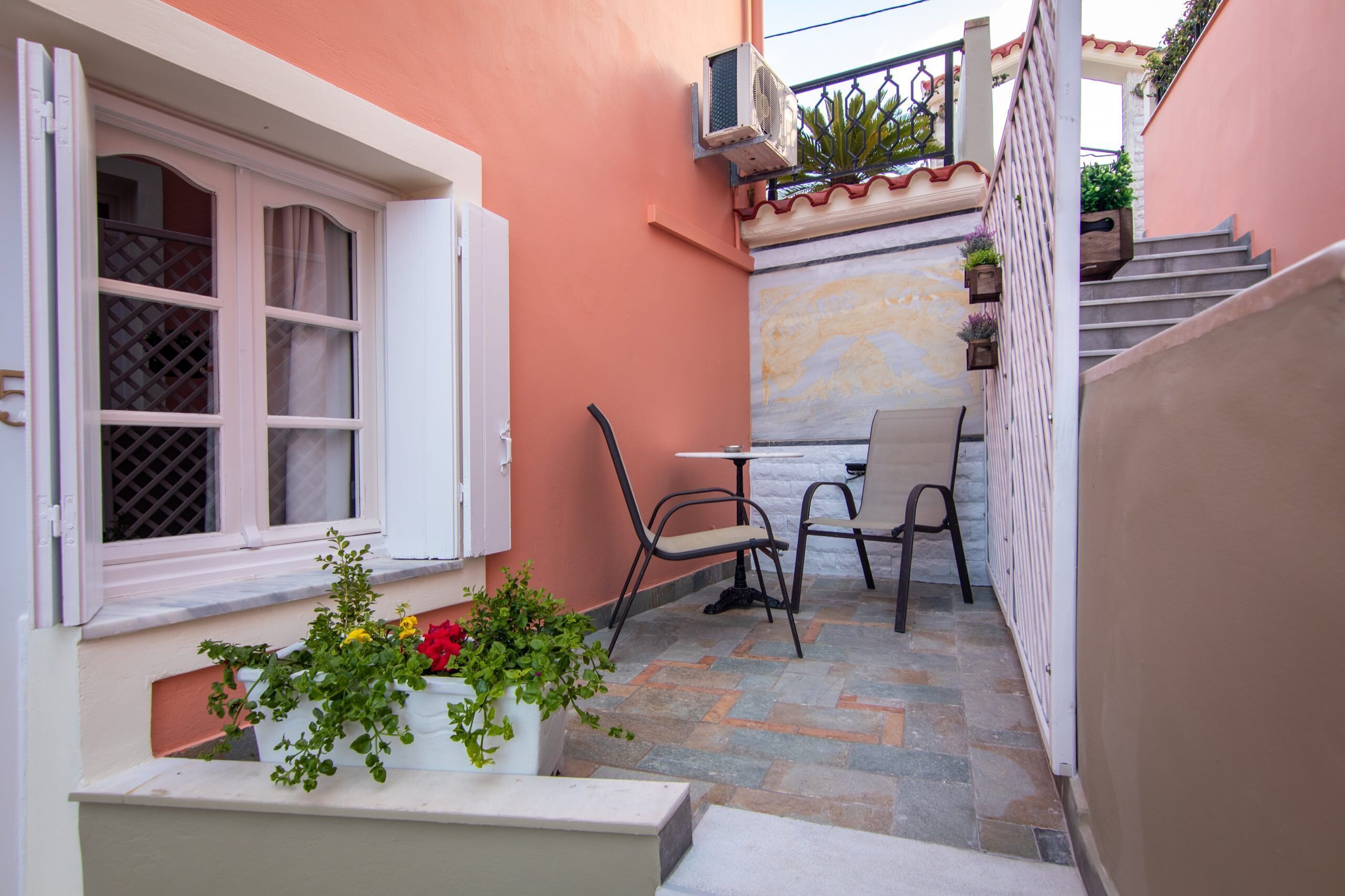 Un piccolo patio esterno con tavolo e sedie, una finestra con persiane e piante in vaso.