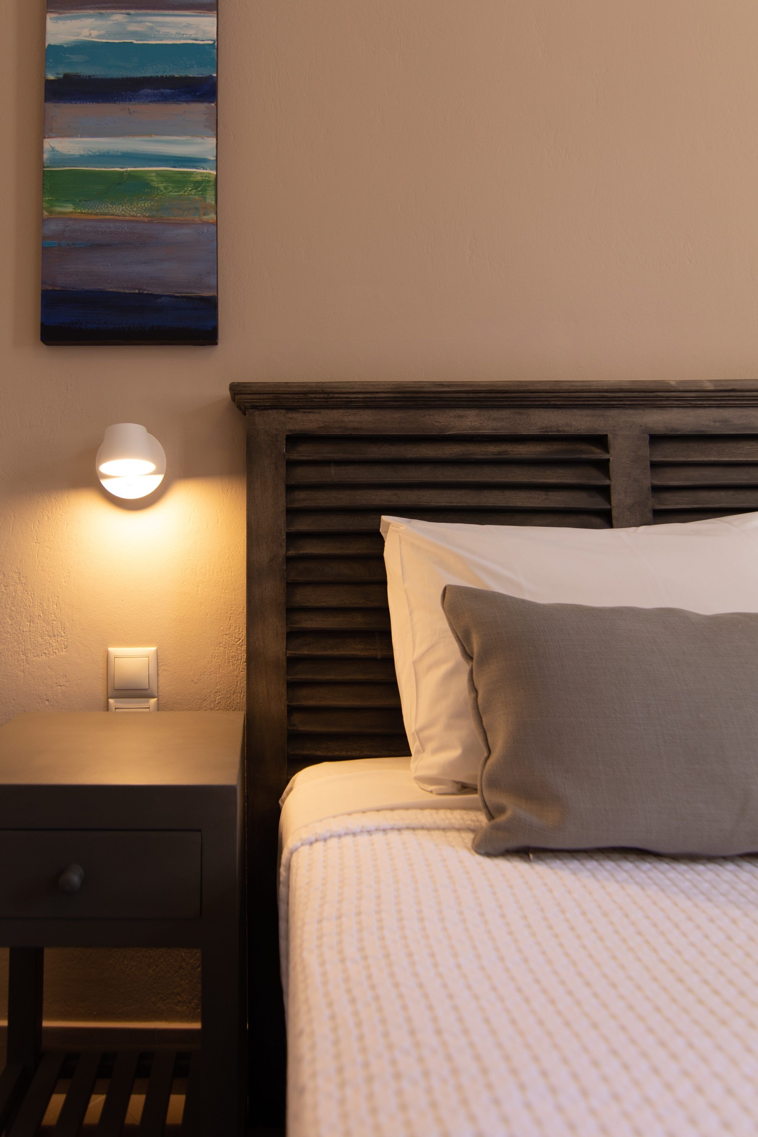 A neatly made bed with a patterned bedspread and pillows, a bedside lamp, and a painting on the wall.