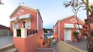 A pink building with a staircase leading to a balcony, a door, and a small garden area. - Fiscardo Studios (Kefalonia)