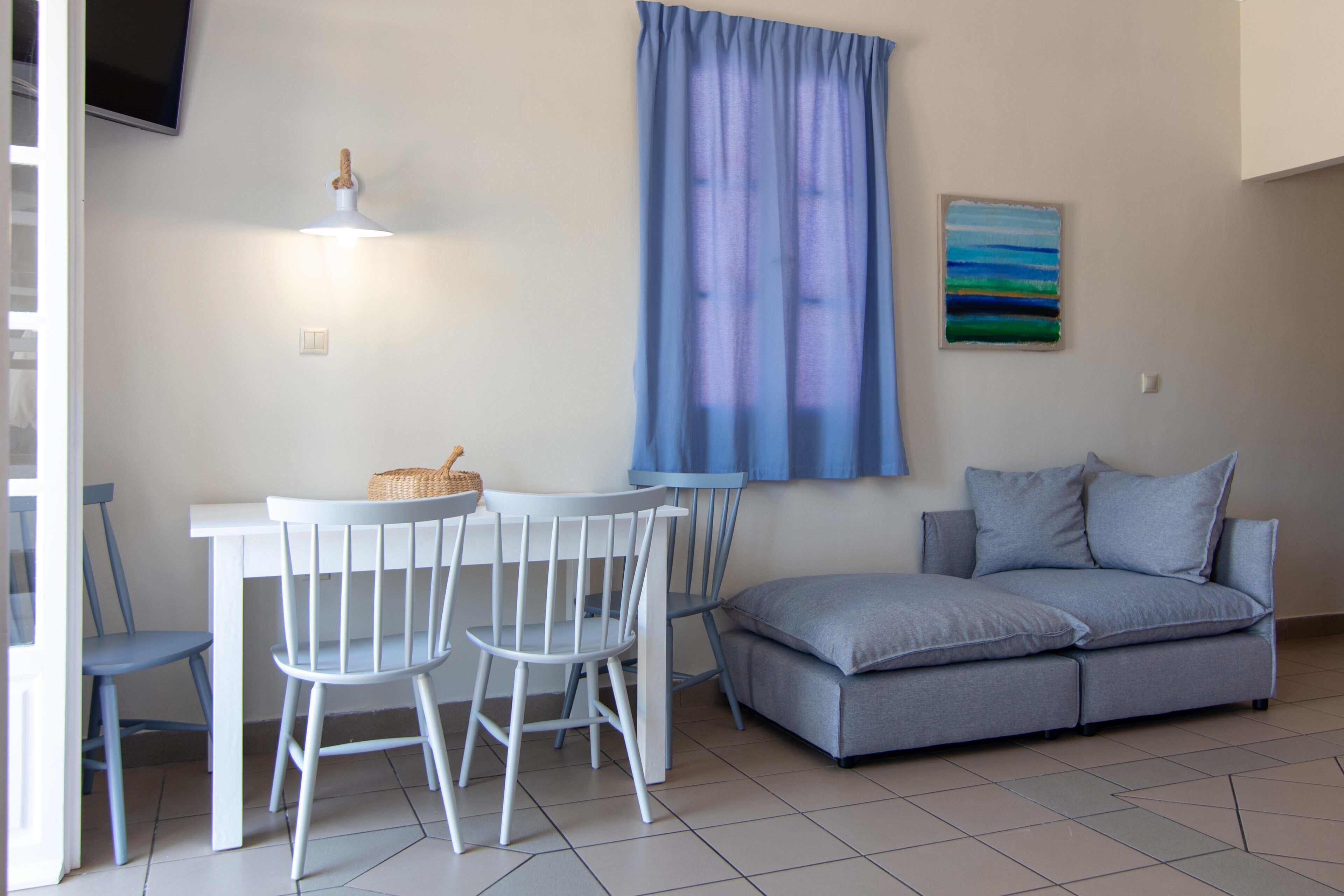 A room with a white table and chairs, a grey sofa, a blue curtain, and a painting on the wall.