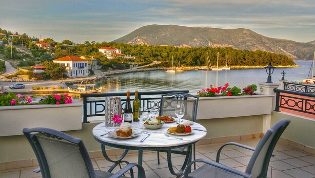 A terrace with a table set for dining, overlooking a body of water with boats and a mountainous backdrop. - Fiscardo Studios (Kefalonia)