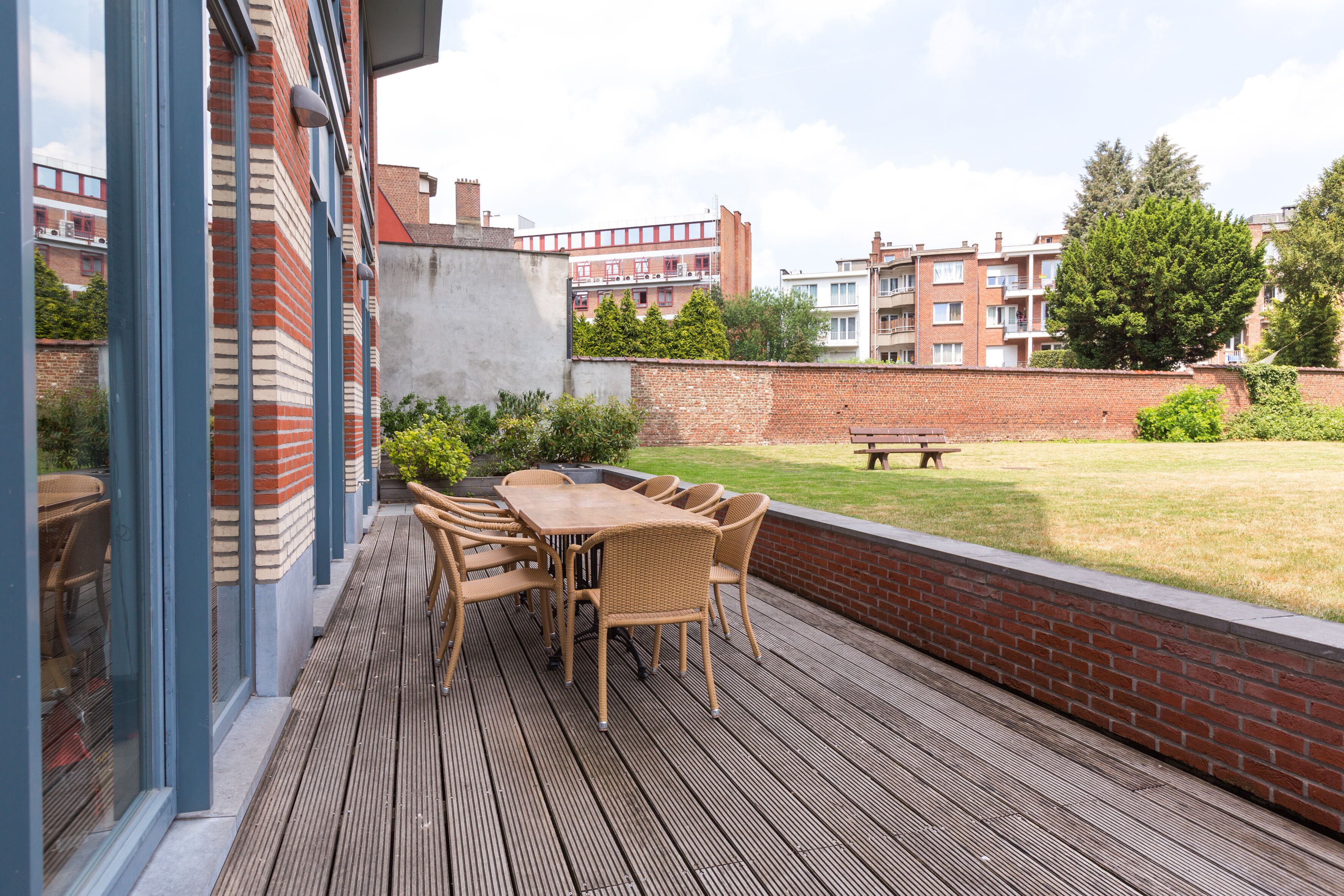 A wooden deck with a table and chairs, a brick wall, and a grassy area with a bench.