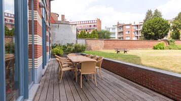A wooden deck with a table and chairs, a brick wall, and a grassy area with a bench.