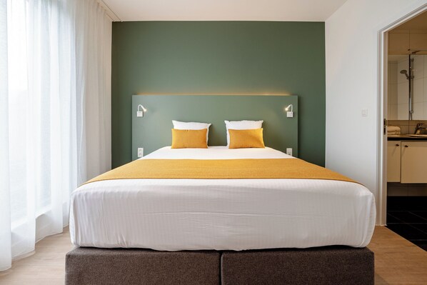 Habitación de hotel con cama, dos lámparas de noche, ventana con cortinas y vista al baño.
