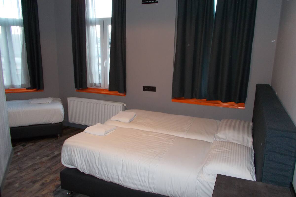 A hotel room with two single beds, a large bed, a wooden floor, a window with curtains, and a radiator.
