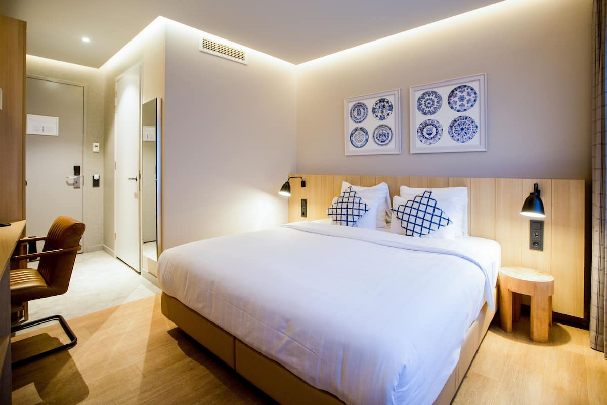 A modern hotel room with a large bed, a wooden chair, a small wooden stool, and wall-mounted artwork.
