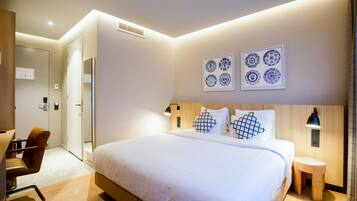 Habitación de hotel moderna con una cama grande, una silla de madera, un taburete pequeño de madera y una obra de arte colgada en la pared.
