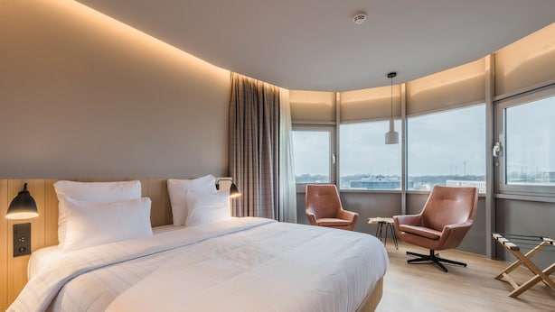 A modern hotel room with a large bed, two armchairs, a small table, and a view of the city through large windows. - Urban Lodge Hotel (Amsterdam)