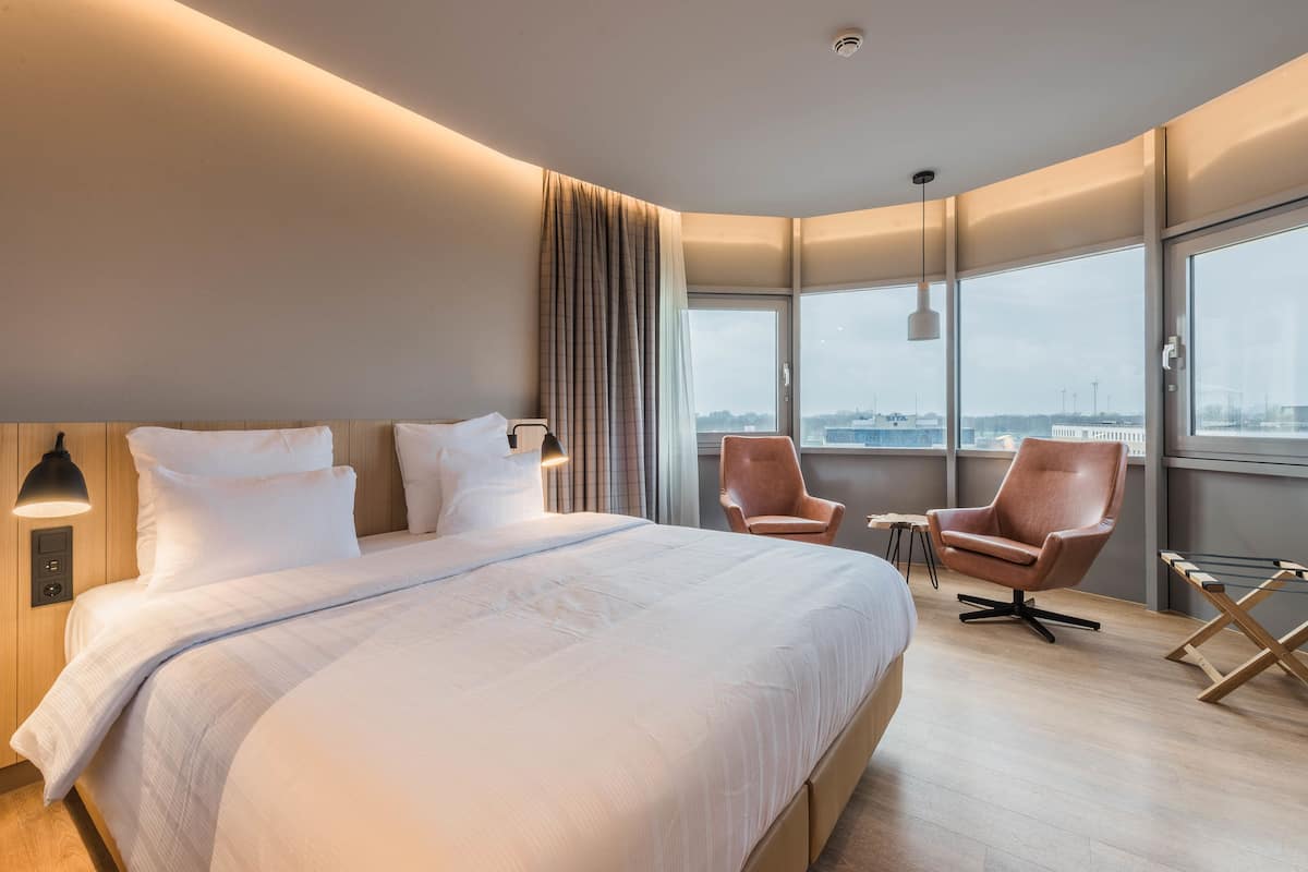 A modern hotel room with a large bed, two armchairs, a small table, and a view of the city through large windows.