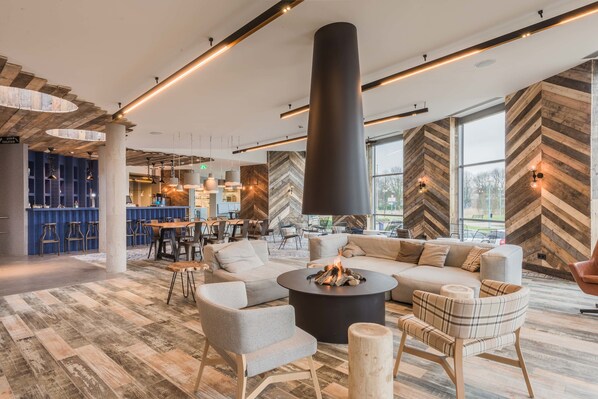 A modern interior with a central seating area, a fireplace, and a bar area in the background. - Urban Lodge Hotel (Amsterdam)