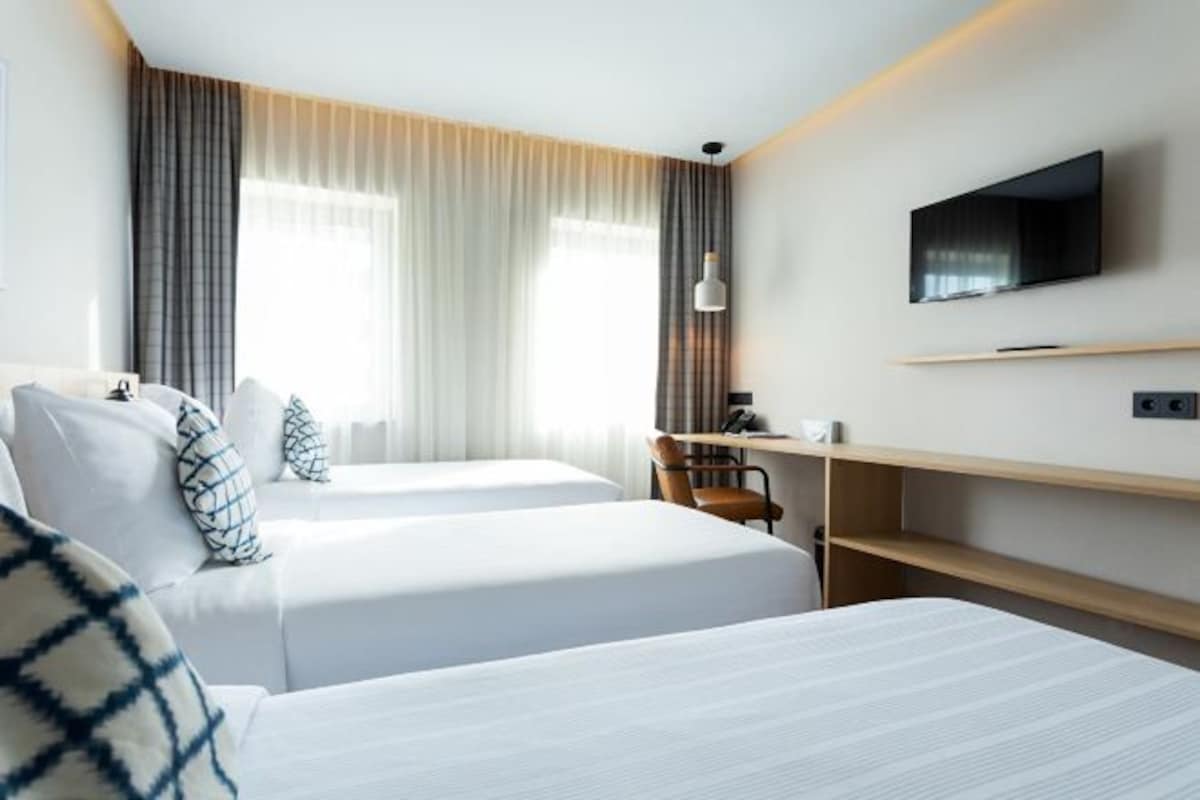 A hotel room with two beds, a desk, a chair, a TV, and a window with curtains.