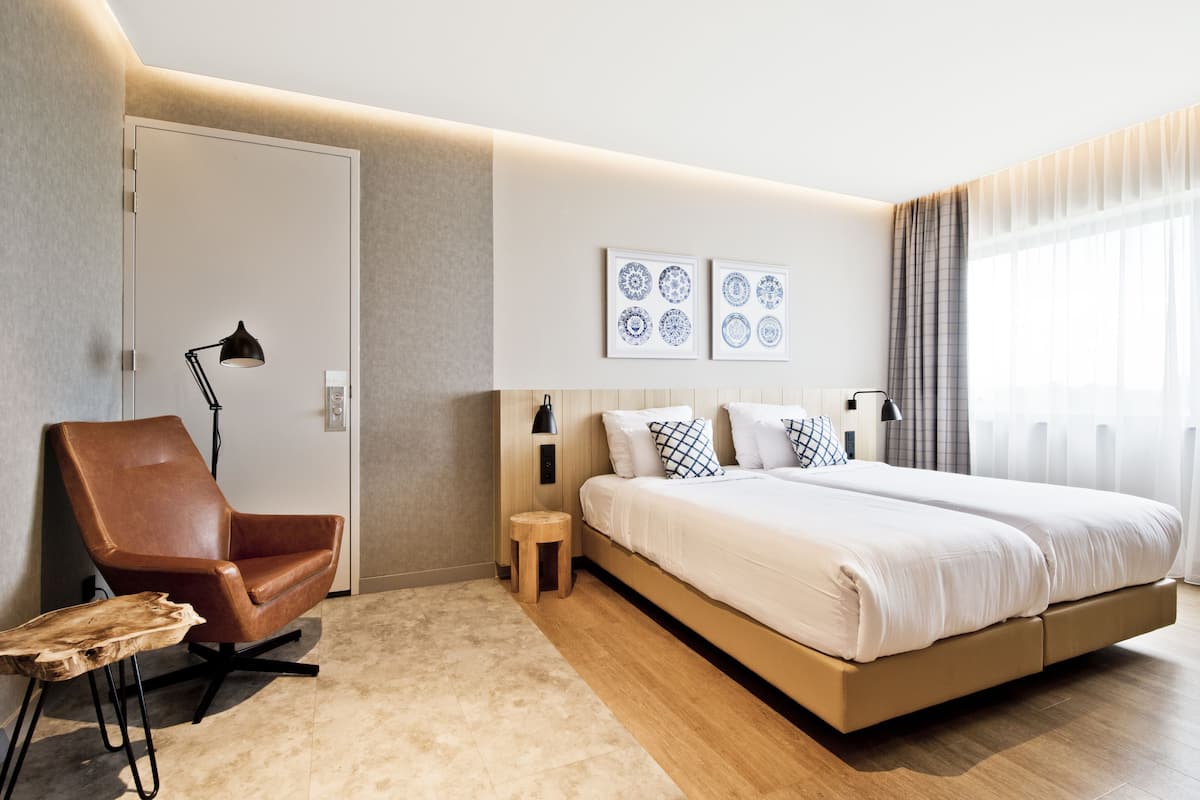 A modern hotel room with a large bed, a brown leather chair, a small wooden stool, and a wall-mounted lamp.