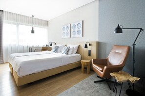 A modern hotel room with a large bed, a brown leather chair, a small wooden stool, and a floor lamp. - Urban Lodge Hotel (Amsterdam)