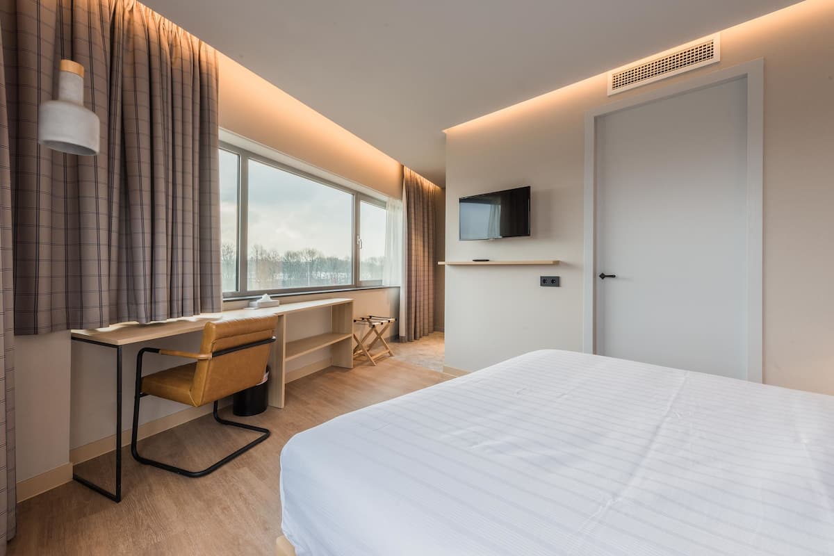 A modern hotel room with a large bed, a desk with a chair, a TV mounted on the wall, and a window with curtains.