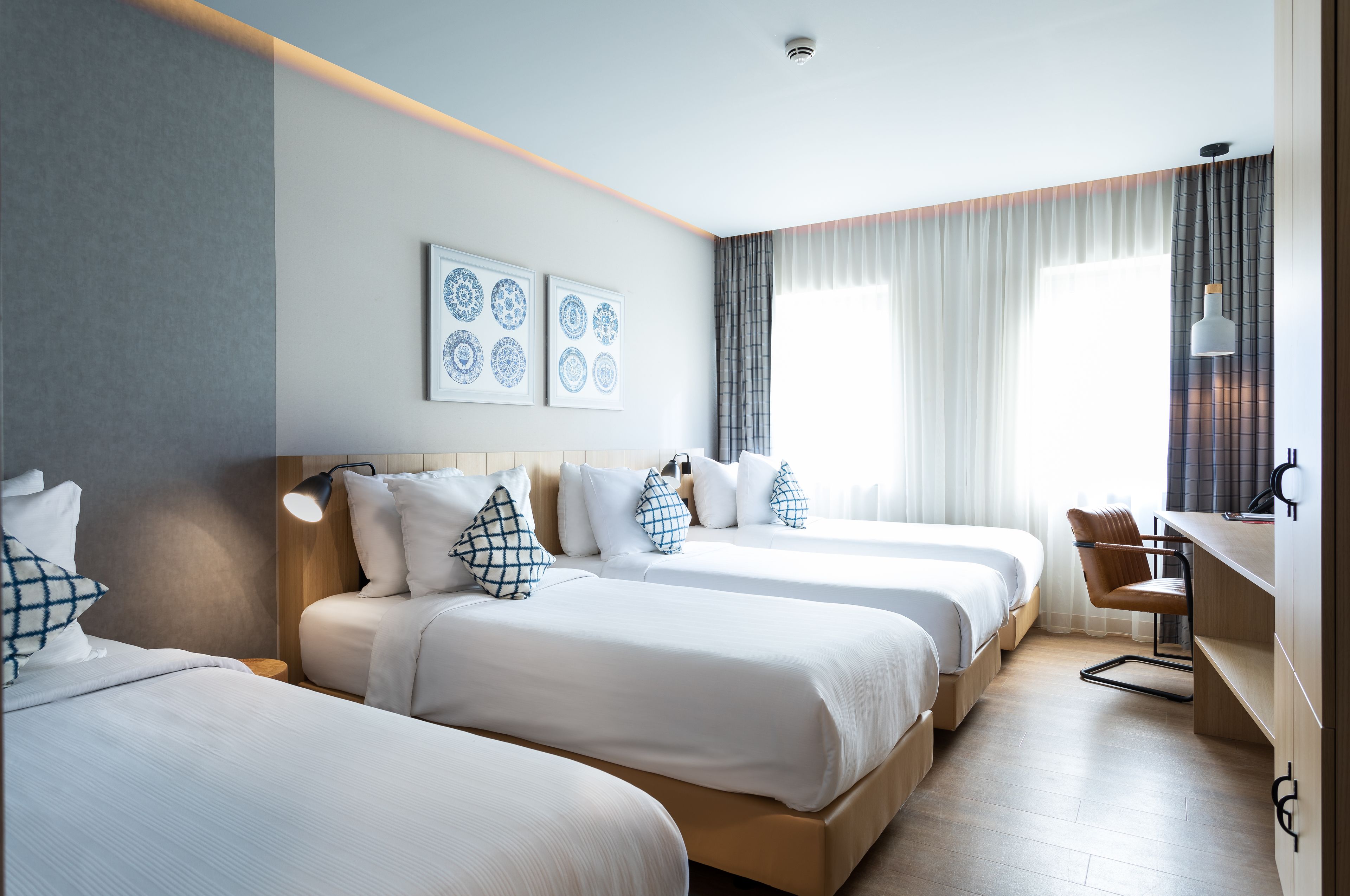 Une chambre d’hôtel avec trois lits, un bureau et une chaise. Il y a deux lampes fixées au mur et un luminaire au plafond.